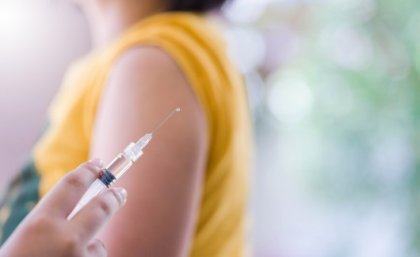 Close-up of vaccine with women in background with yellow shirt sleeves rolled up.